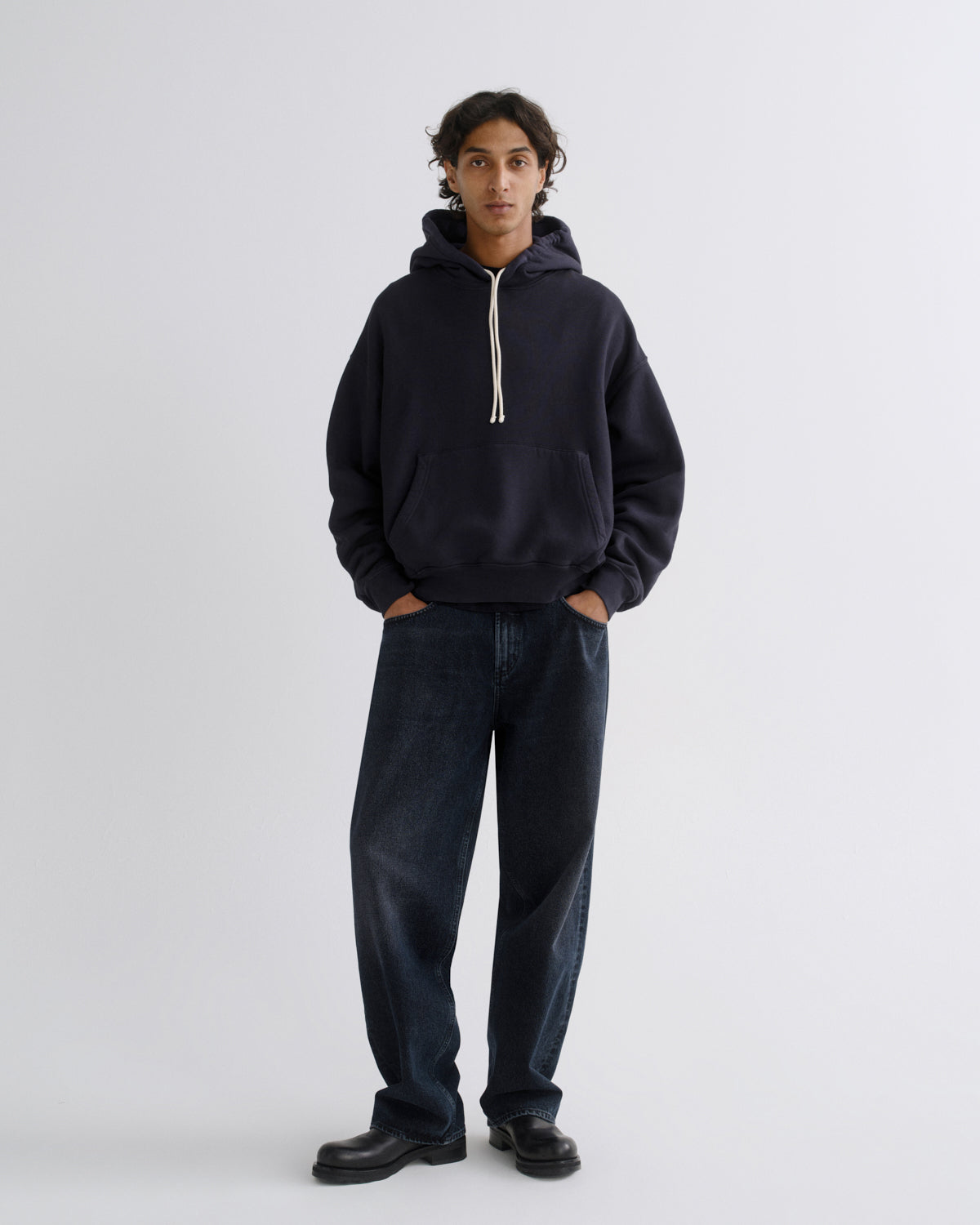 A model stands against a light gray studio background wearing a dark navy pullover hoodie with cream drawstrings, dark blue jeans, and black leather shoes.