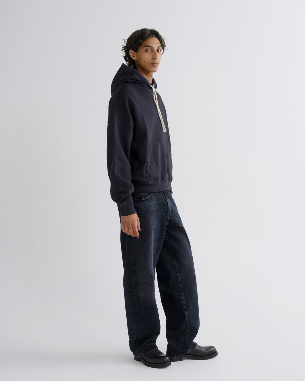 A man wearing a dark navy blue pullover hoodie with beige drawstrings and dark blue jeans, standing in profile against a light gray studio background.
