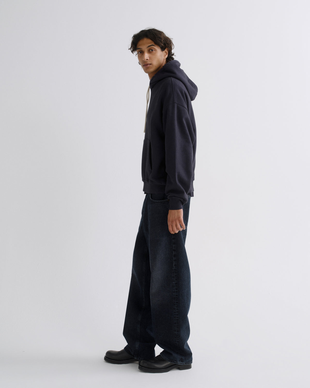 A model wearing a dark navy blue hoodie and matching dark wide-leg jeans, standing in profile against a light gray studio background.