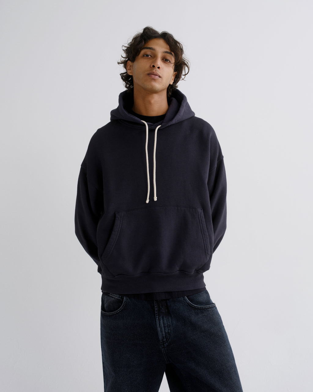 A man wearing a dark navy pullover hoodie with cream-colored drawstrings and dark blue jeans, standing against a light gray studio background.