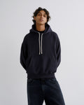 A man wearing a dark navy pullover hoodie with cream-colored drawstrings and dark blue jeans, standing against a light gray studio background.
