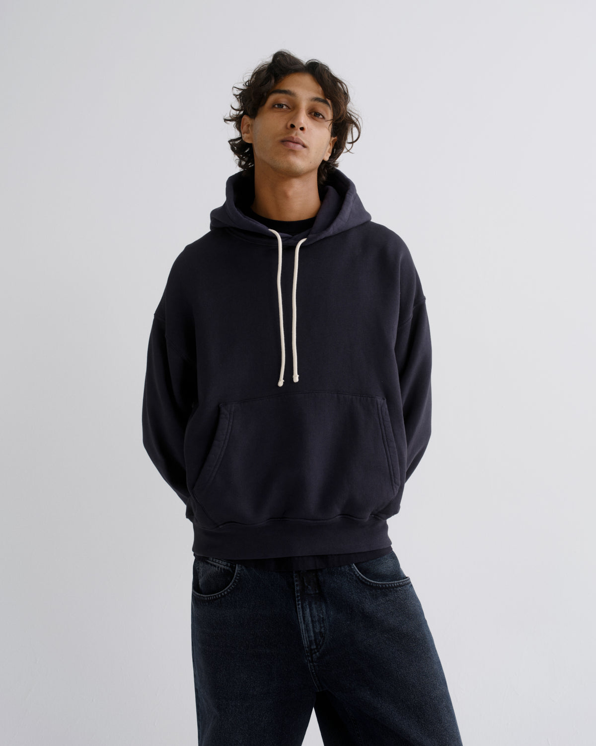 A man wearing a dark navy pullover hoodie with cream-colored drawstrings and dark blue jeans, standing against a light gray studio background.