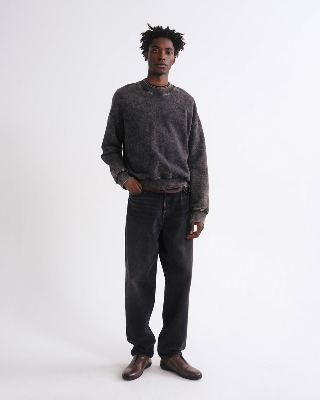 A man modeling a dark gray washed crewneck sweatshirt and dark gray jeans with brown leather shoes, standing against a plain white studio background.
