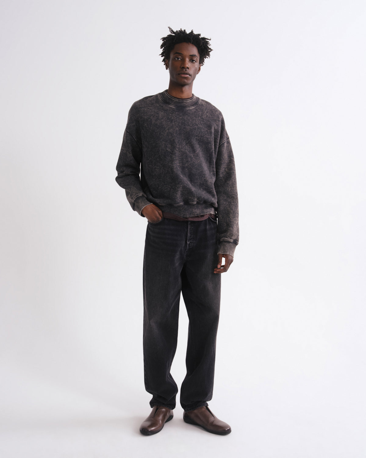 A man modeling a dark gray washed crewneck sweatshirt and dark gray jeans with brown leather shoes, standing against a plain white studio background.