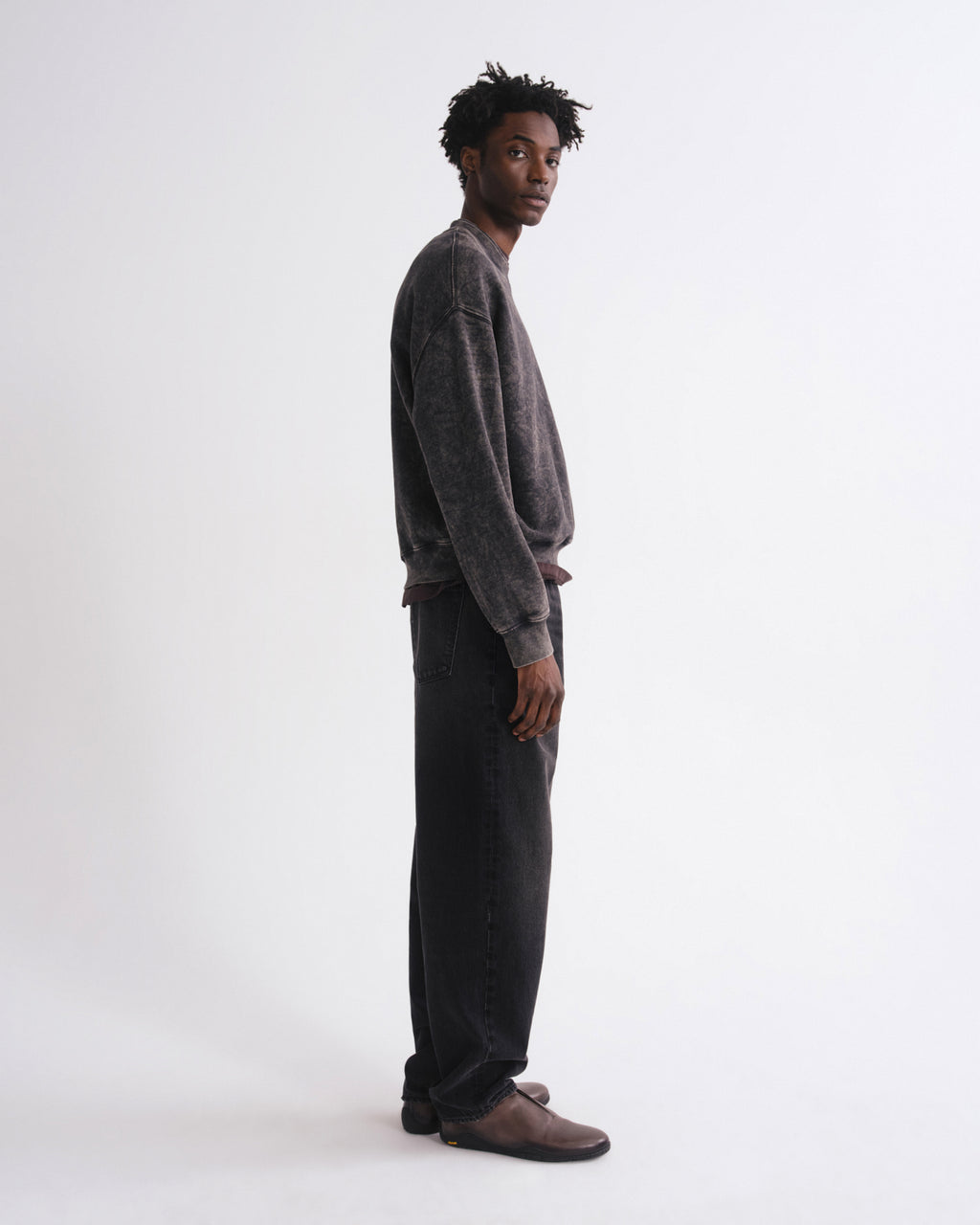 A man stands in profile wearing a dark gray washed crewneck sweatshirt, dark charcoal pants, and brown suede loafers against a plain white studio background.