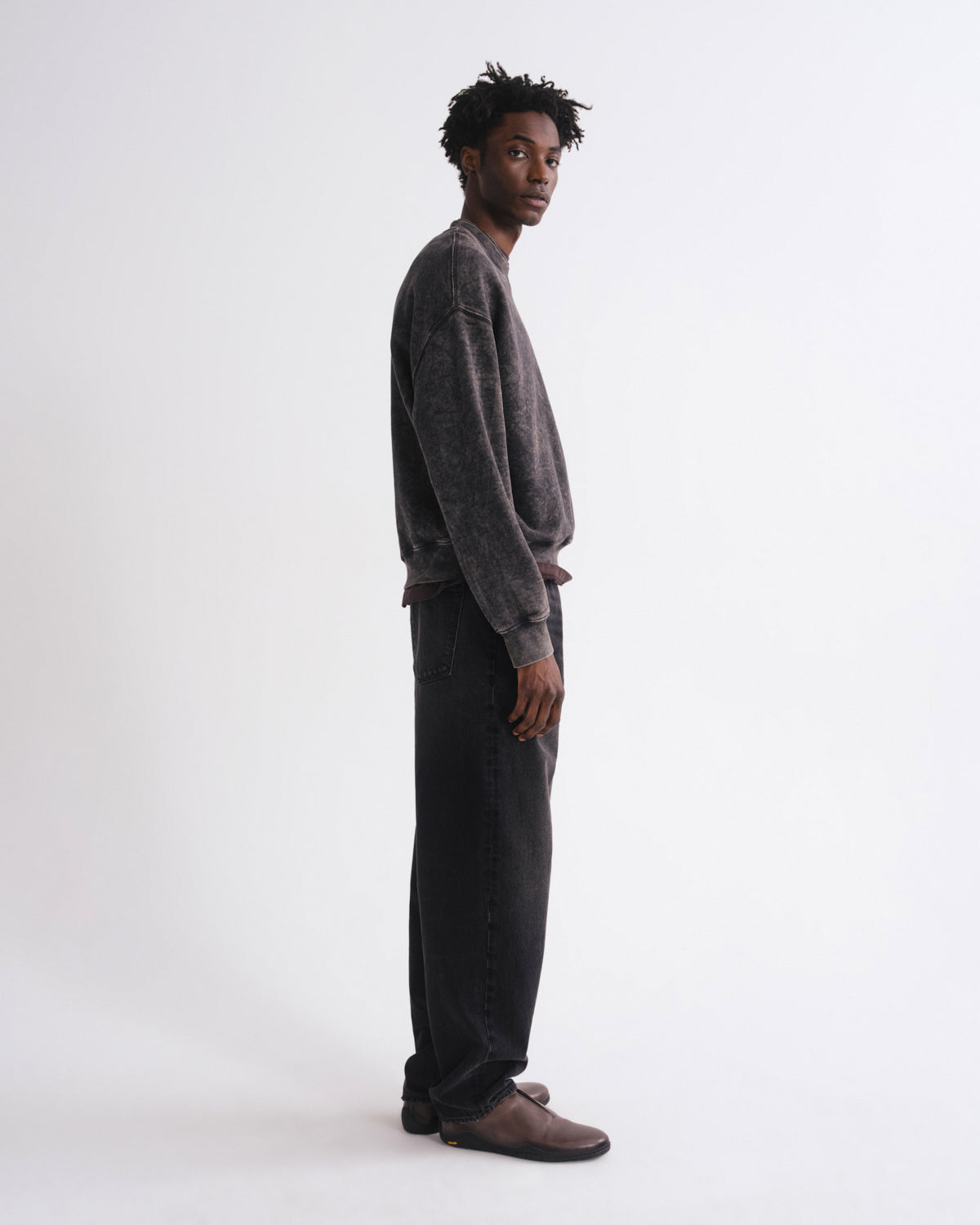 A man stands in profile wearing a dark gray washed crewneck sweatshirt, dark charcoal pants, and brown suede loafers against a plain white studio background.