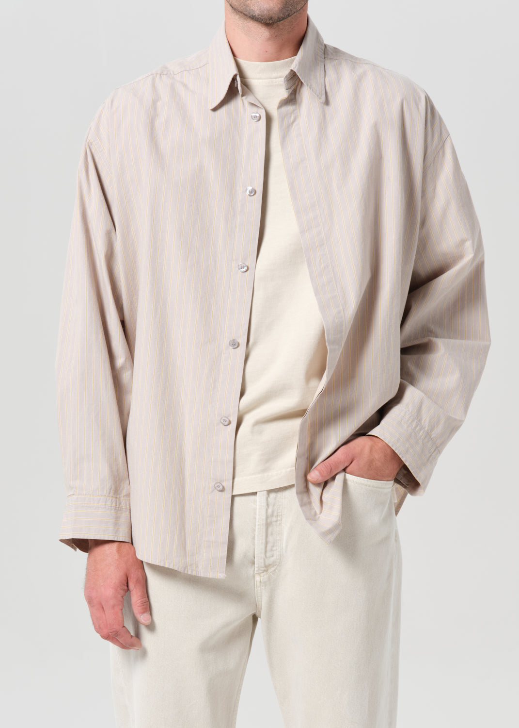 Person wearing a beige button-up shirt over a white shirt with light-colored pants on a plain background