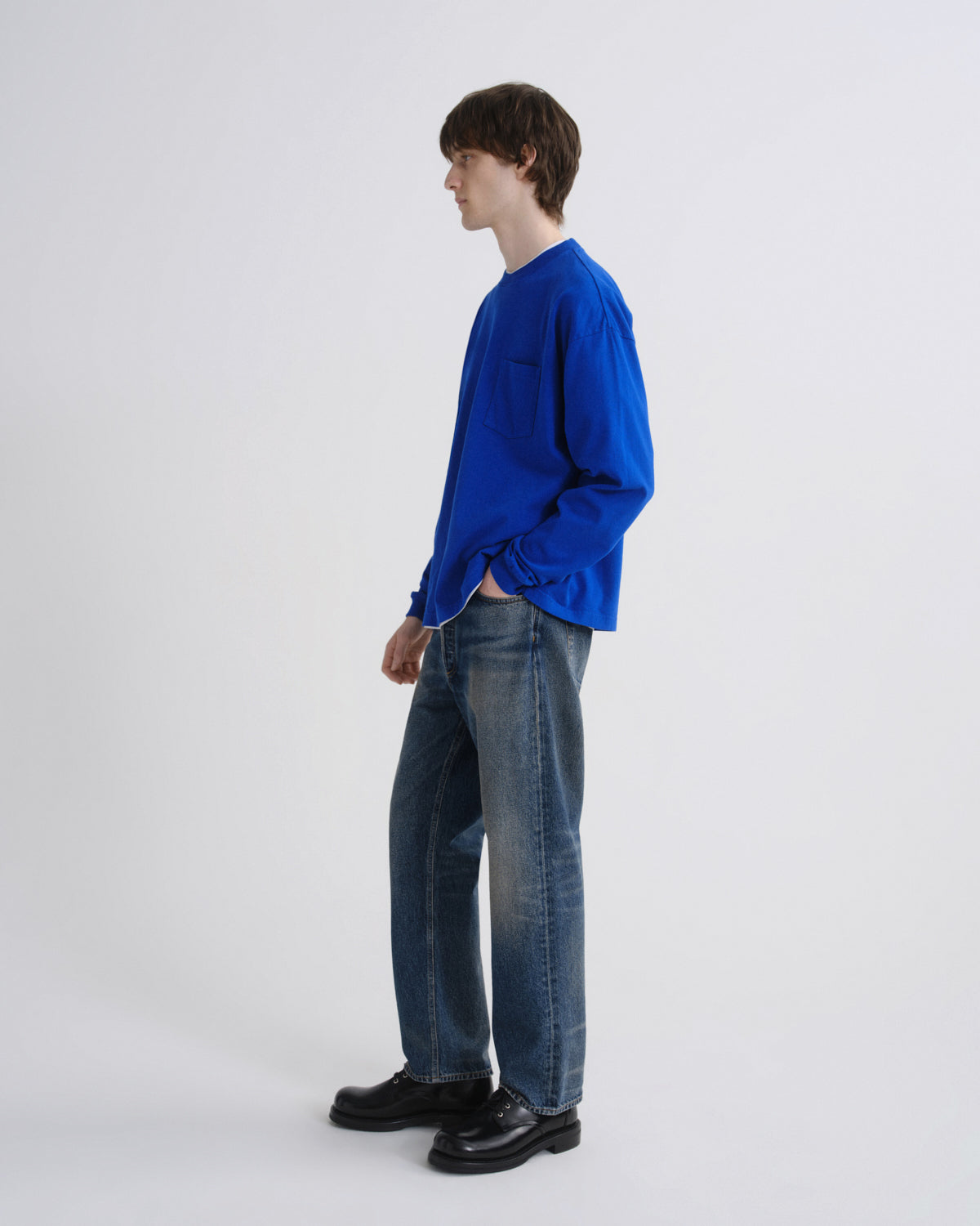 Person wearing a blue shirt and dark jeans standing against a grey background