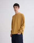 A man wearing a mustard yellow long-sleeve crewneck shirt with a chest pocket, paired with dark blue jeans, standing against a light gray studio background.