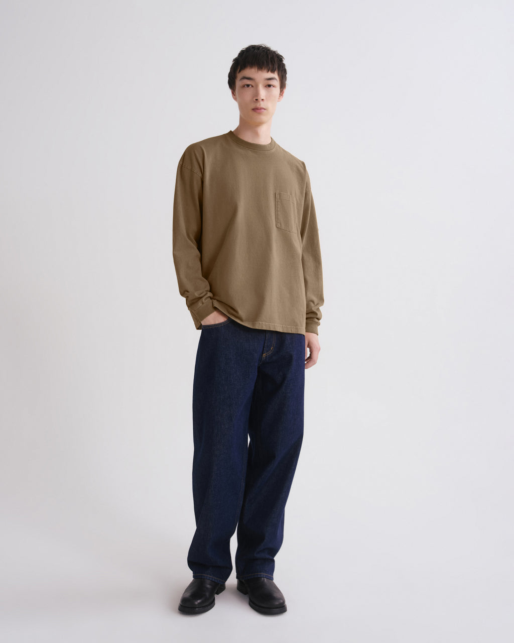 A man modeling a long-sleeve crewneck shirt in a muted olive-brown color, paired with dark blue jeans and black shoes, standing against a plain light gray studio background.
