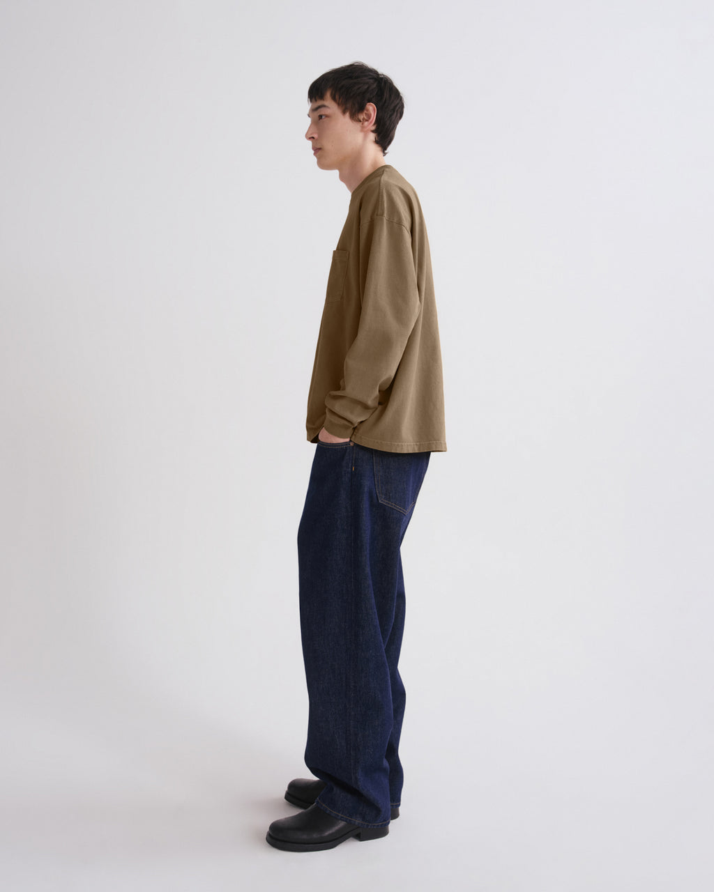 A man stands in profile wearing a loose olive-brown long-sleeve shirt, dark blue wide-leg jeans, and black shoes against a plain light gray studio background.
