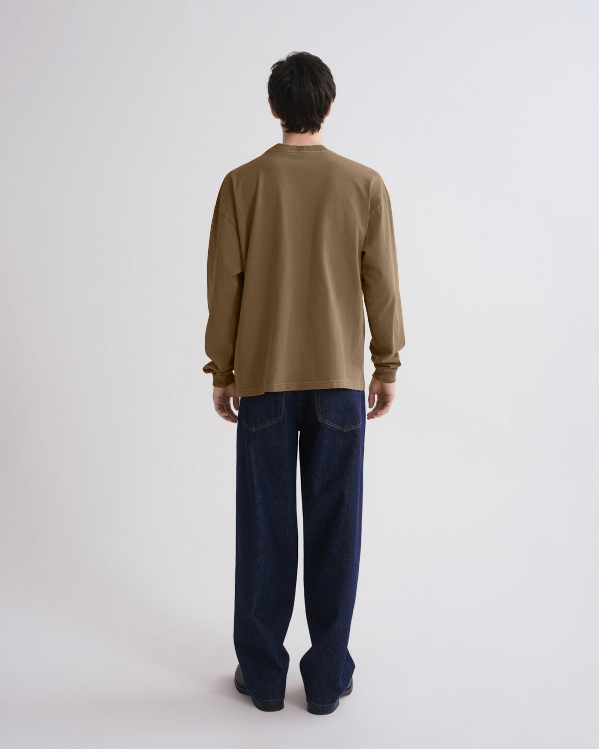A man viewed from behind wearing a muted brown long-sleeve shirt and dark blue jeans, standing against a plain light gray studio background.