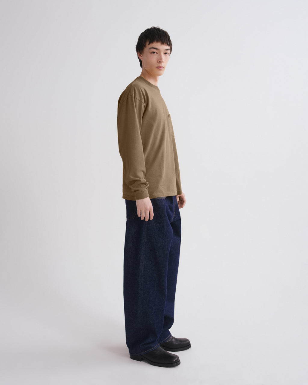 A man stands in profile wearing a loose olive-brown long-sleeve shirt, dark blue wide-leg jeans, and black shoes against a plain light gray studio background.