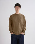A man wearing a muted brown long-sleeve crewneck shirt with a chest pocket and dark blue jeans, standing against a light gray studio background.