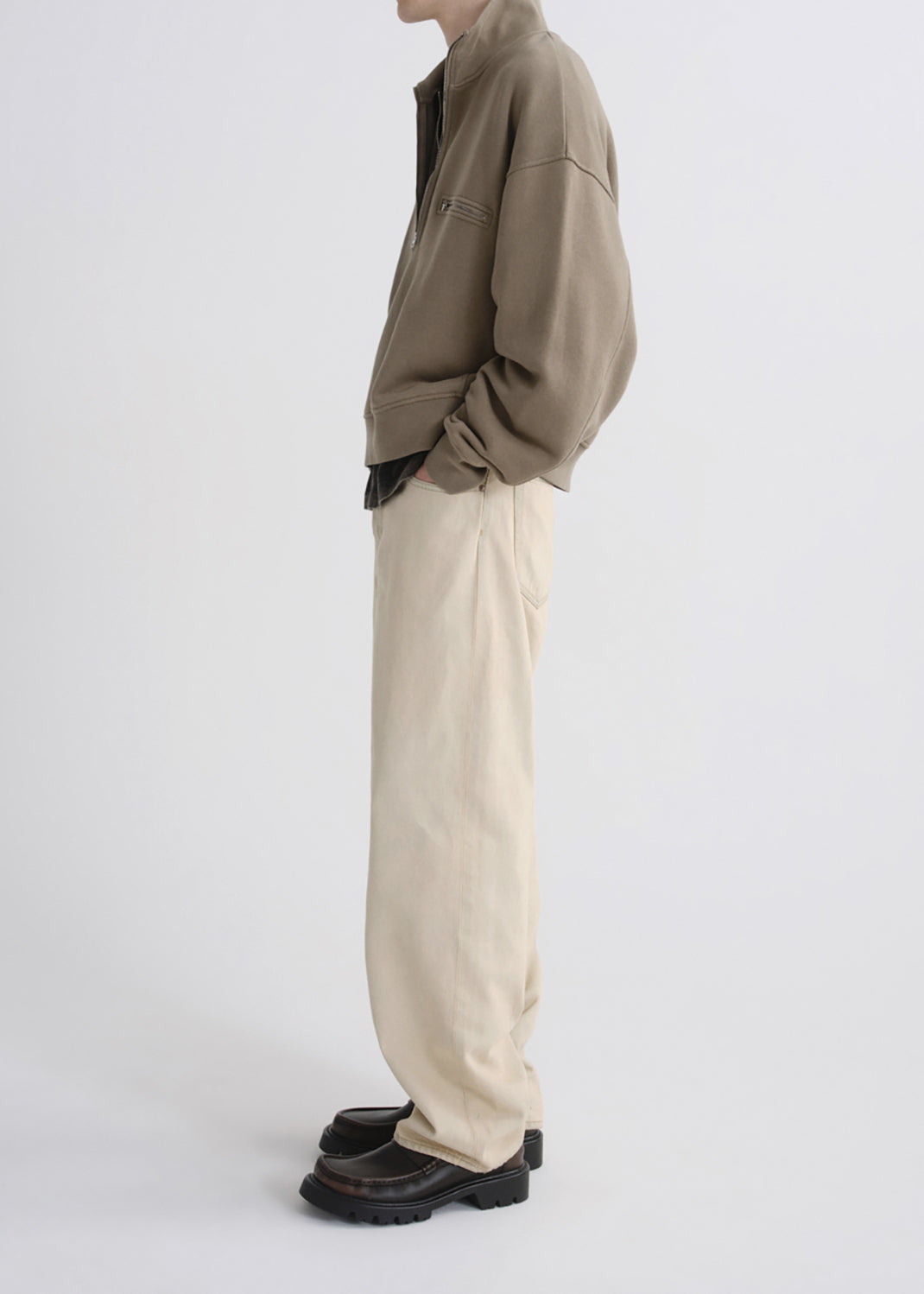 A side-profile studio shot of a model wearing a taupe-brown utility jacket, cream-colored wide-leg trousers, and dark brown leather loafers against a grey background.