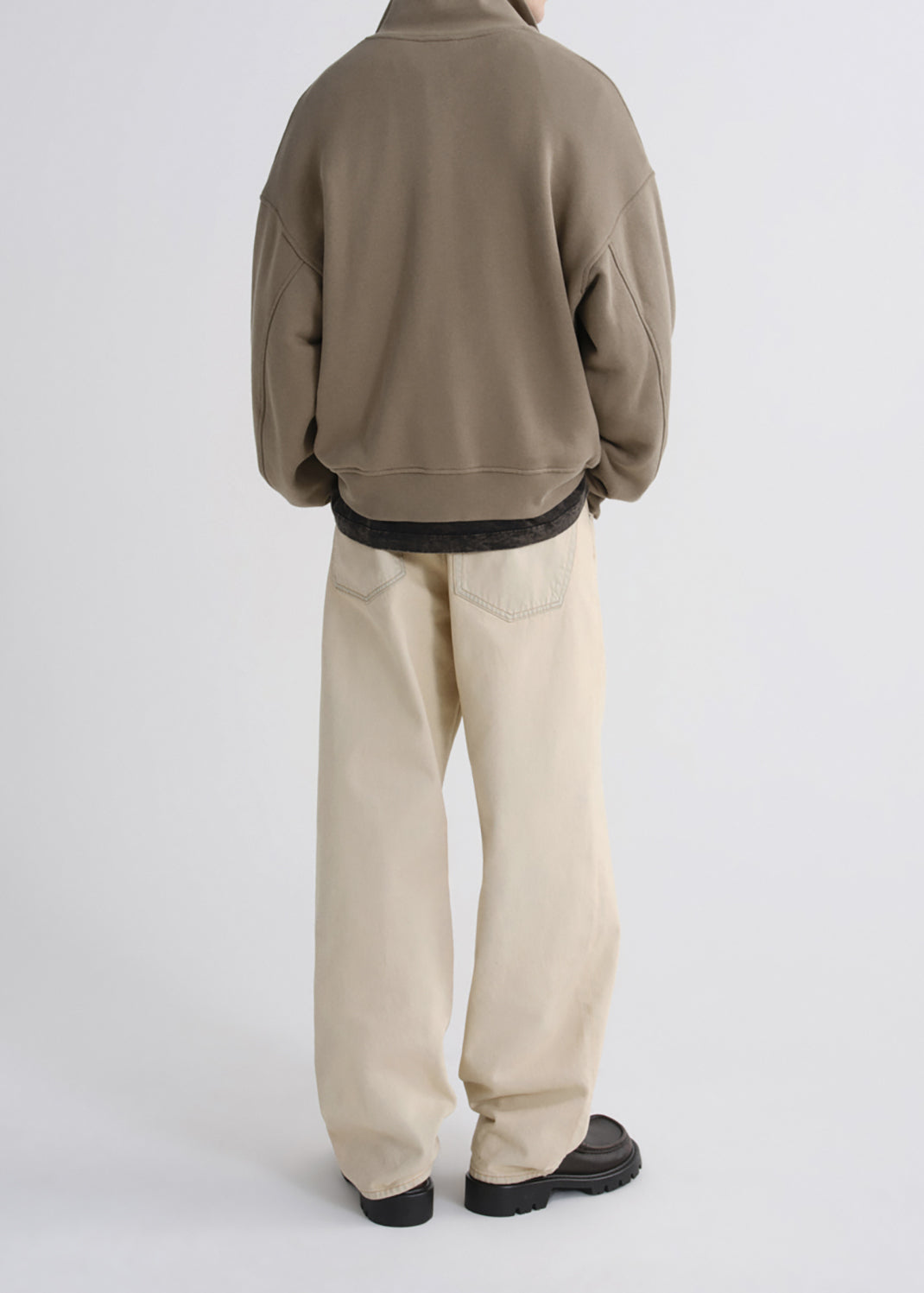 A person shown from the back wearing a taupe-brown cropped sweatshirt, cream-colored wide-leg trousers, and black shoes, photographed against a grey background.
