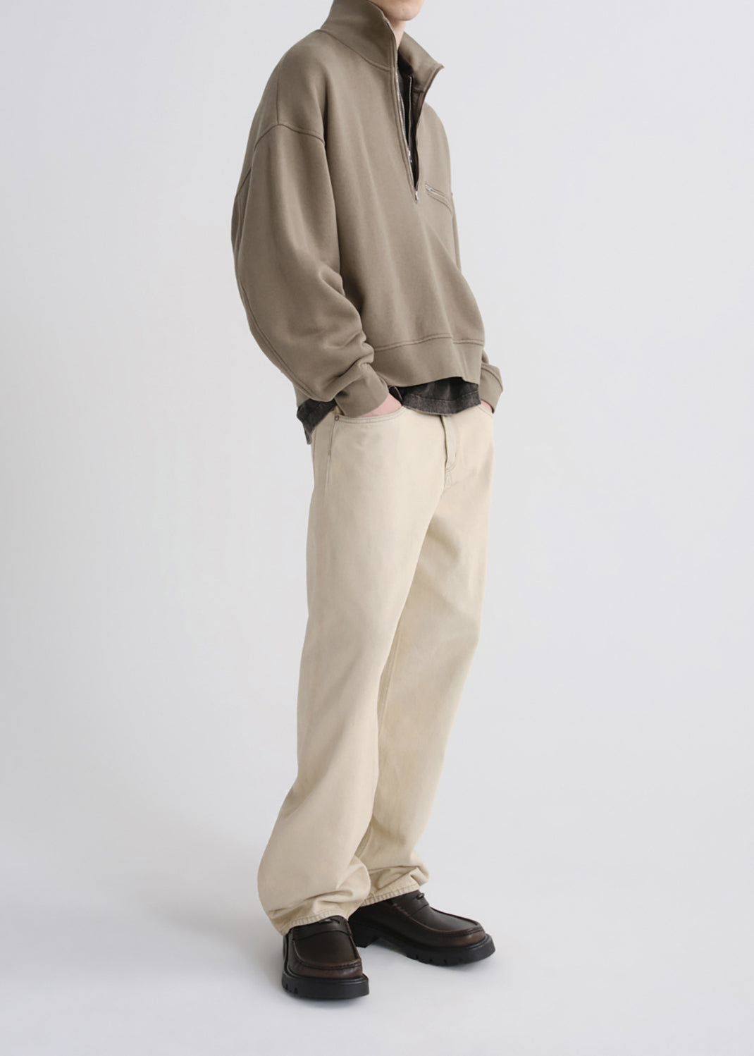 A model wearing a taupe quarter-zip sweatshirt, cream-colored relaxed-fit trousers, and dark brown leather loafers, shown from the neck down against a grey background.