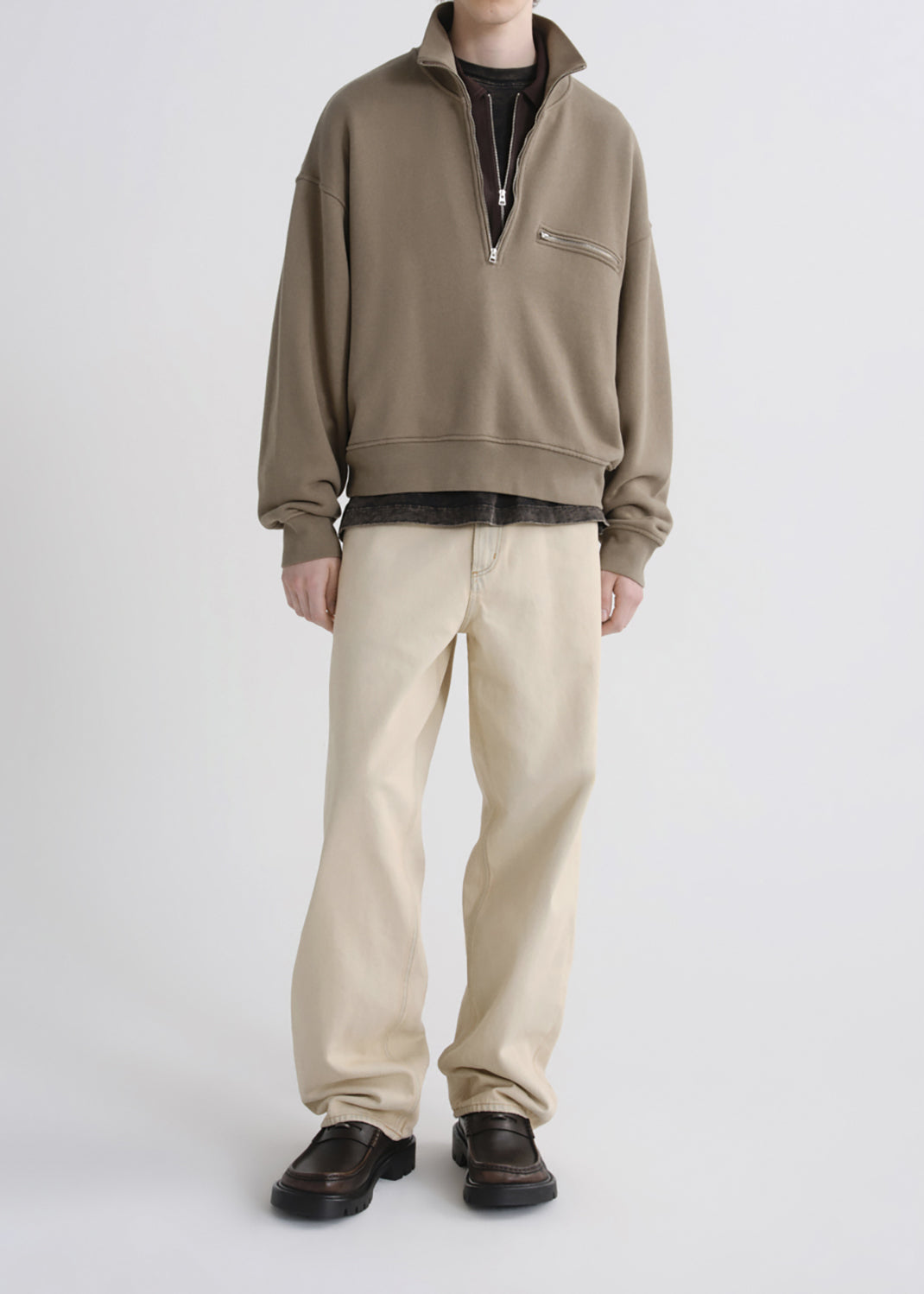 A model wearing a taupe-brown half-zip pullover sweatshirt, beige trousers, and dark brown leather shoes, standing against a grey background.