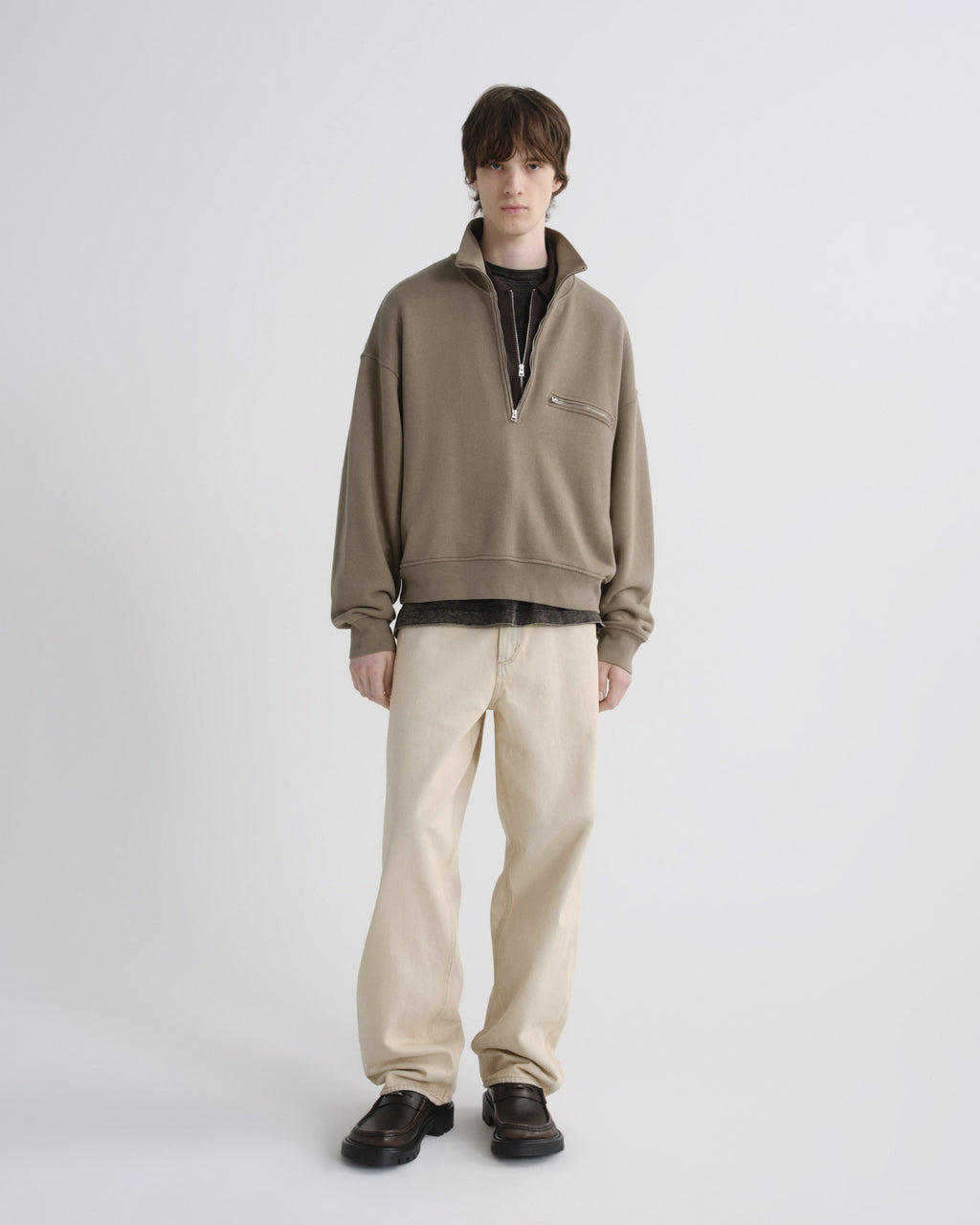 Man wearing a beige pullover with a black shirt underneath, beige pants, and brown shoes on a white background