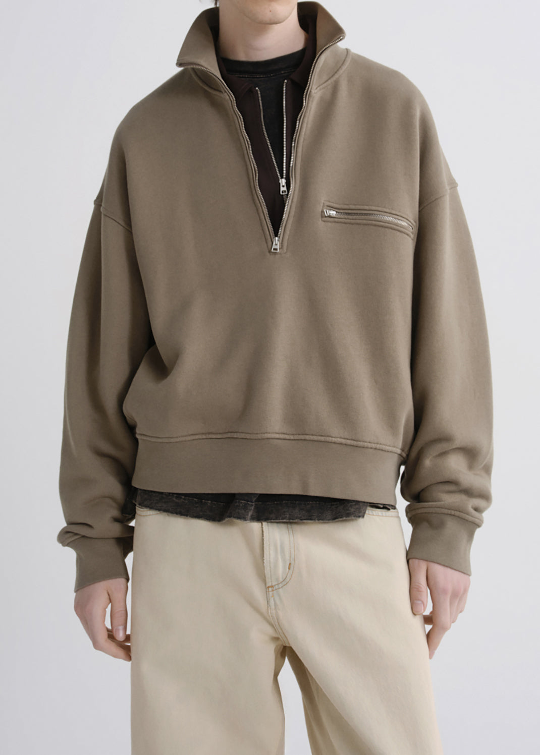 A person wearing an oversized taupe quarter-zip pullover with a zippered chest pocket, layered over a black shirt and paired with beige pants, photographed against a grey background.