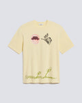 A pale yellow short-sleeve t-shirt with graphic prints, featuring a mouth graphic and the word 'MUTUAL' on the chest, and green script text near the hem, displayed on a light gray background.