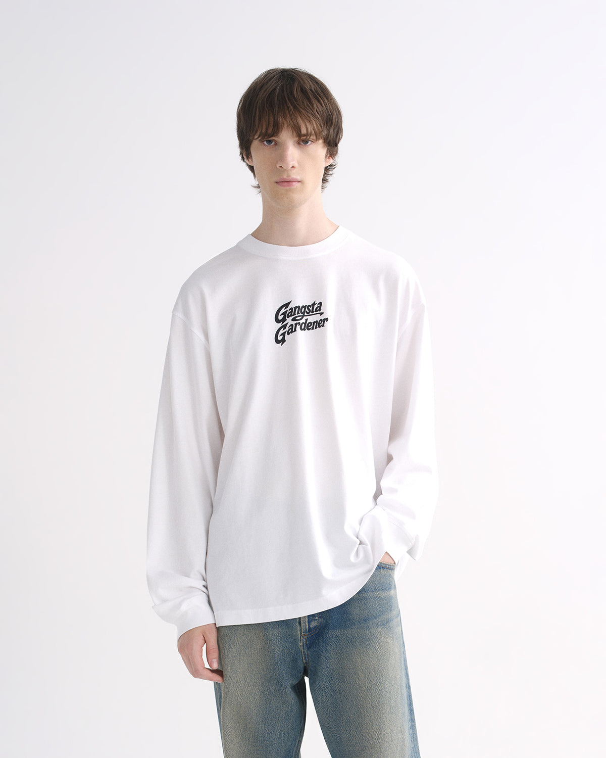 Person wearing a white long sleeve graphic T-shirt with blue jeans and black shoes on a grey background