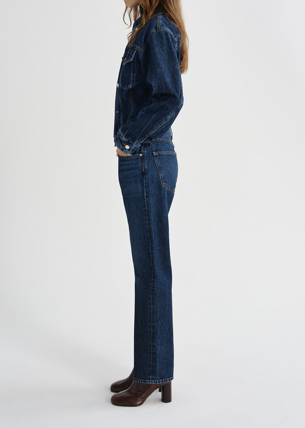 Person wearing a dark wash denim jacket with matching dark wash straight leg jeans viewed from the side on a grey background