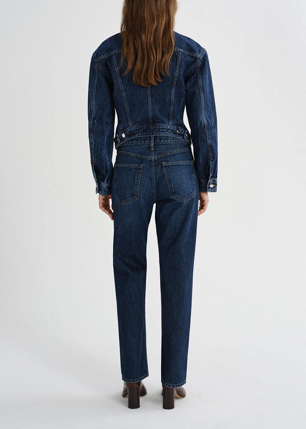 Person wearing a dark wash denim jacket with matching dark wash straight leg jeans viewed from the back on a grey background
