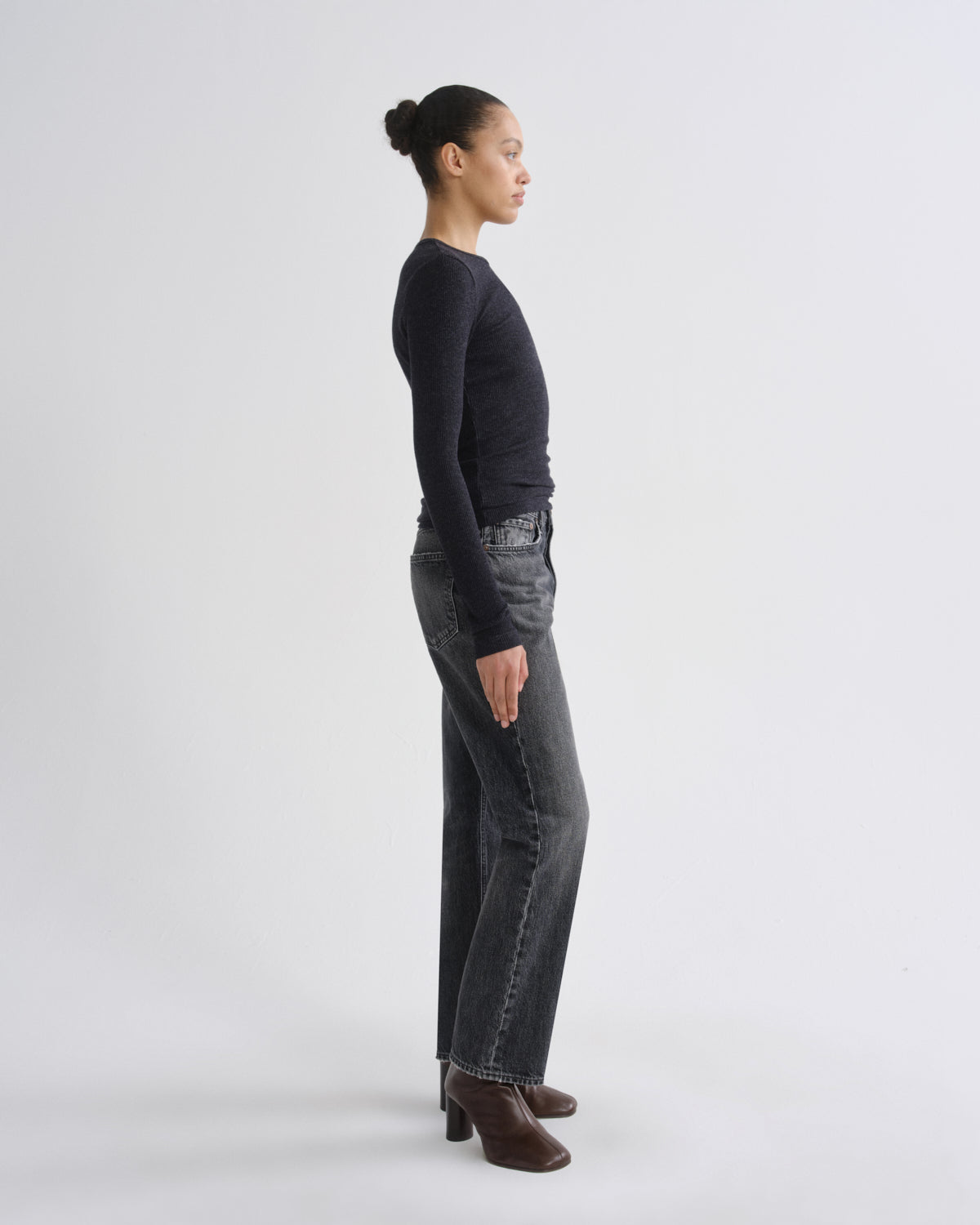 A model shown in profile wearing a dark charcoal long-sleeve knit top, dark gray washed jeans, and brown ankle boots, standing against a light gray studio background.