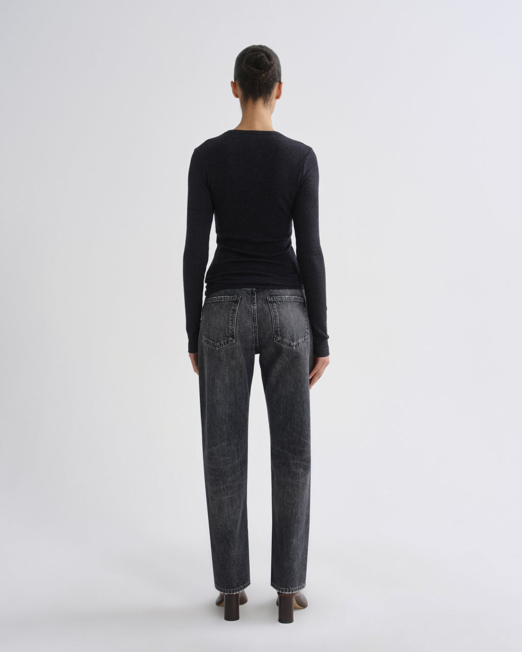 A woman viewed from behind wearing a black long-sleeve top and dark gray washed jeans, standing against a plain light gray studio background.