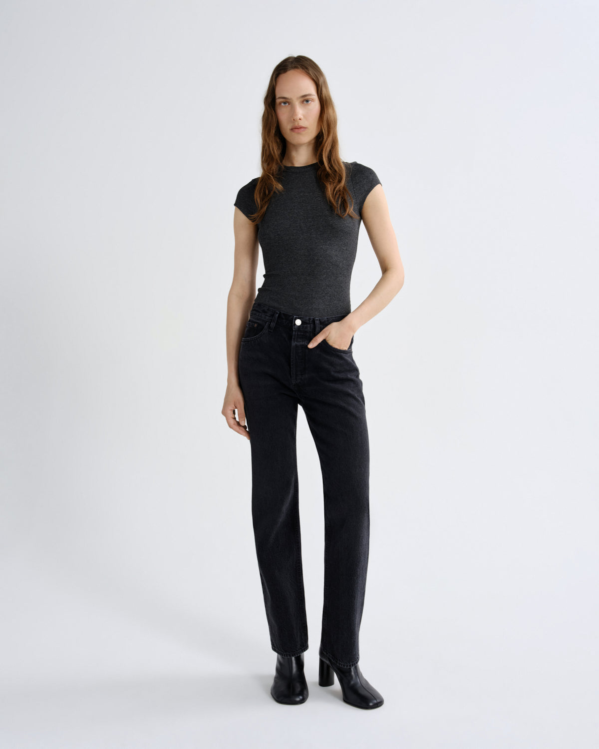 A woman modeling a dark gray short-sleeve knit top and black straight-leg jeans with black ankle boots, standing against a light gray studio background.