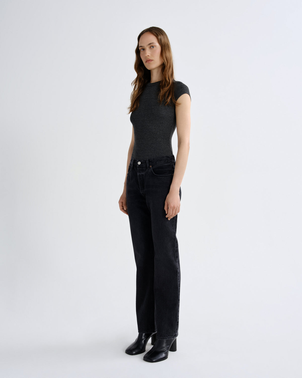 A woman modeling a dark charcoal gray sleeveless top and black wide-leg jeans with black ankle boots, standing against a plain light gray studio background.