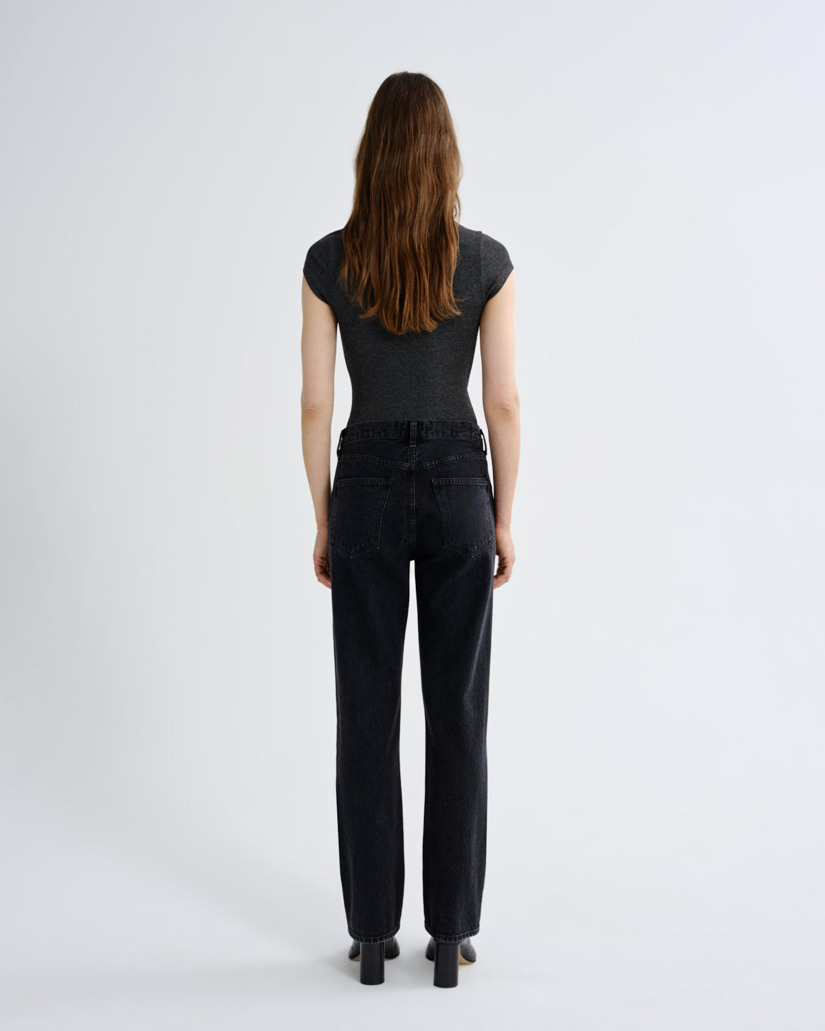 A woman viewed from behind wearing a dark gray short-sleeve top and black wide-leg jeans, standing against a plain light gray studio background.