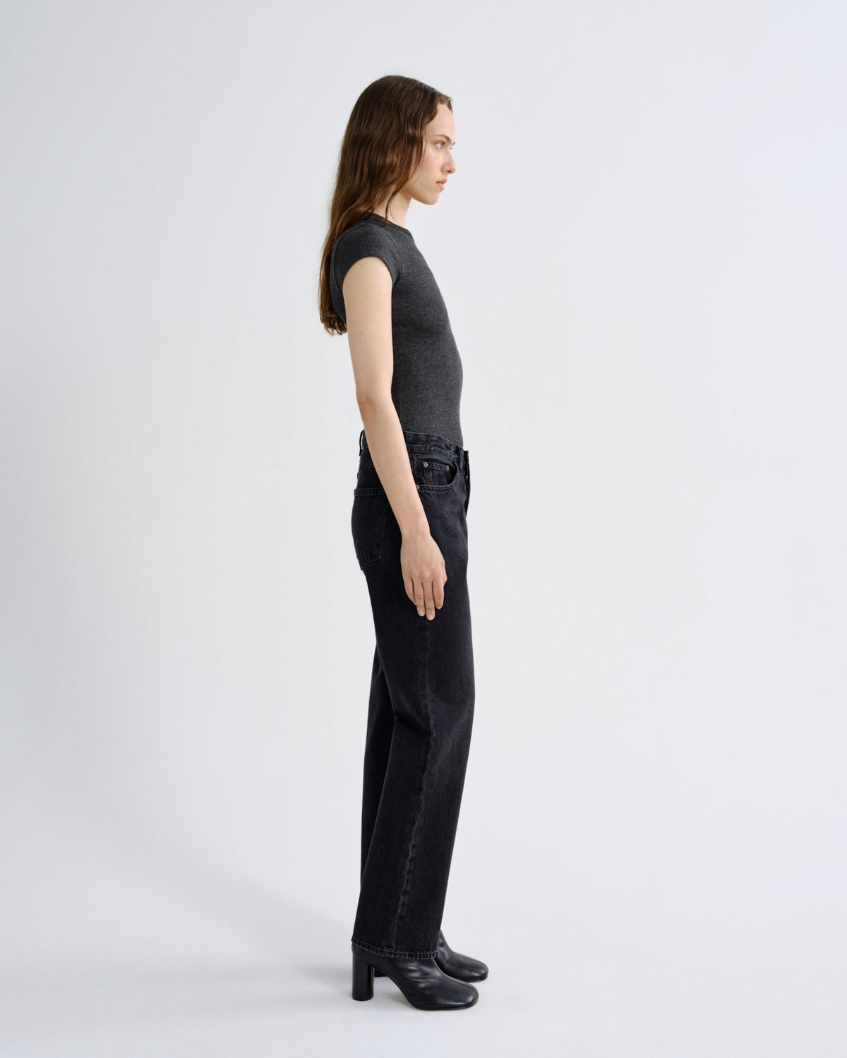 A woman stands in profile wearing a fitted dark charcoal sleeveless top, black wide-leg jeans, and black heeled boots against a clean light gray studio background.