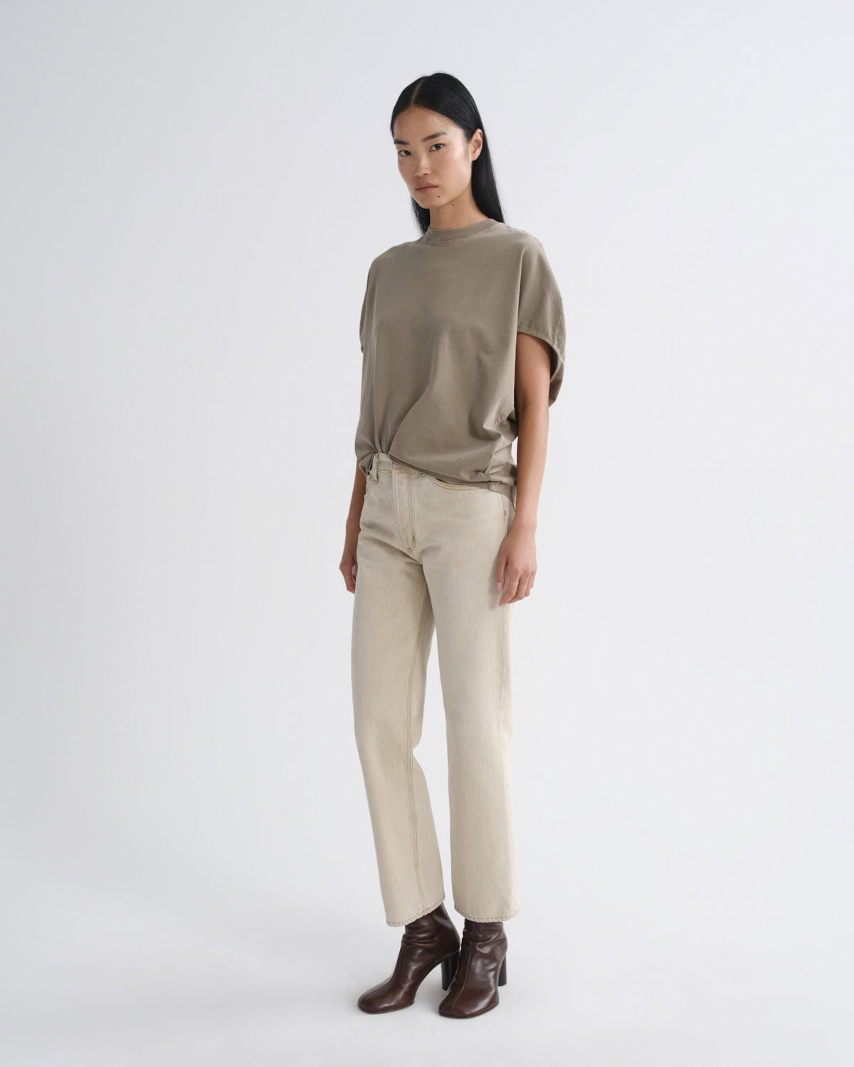 A woman modeling a loose taupe short-sleeve top, beige straight-leg trousers, and dark brown ankle boots against a plain light gray studio background.