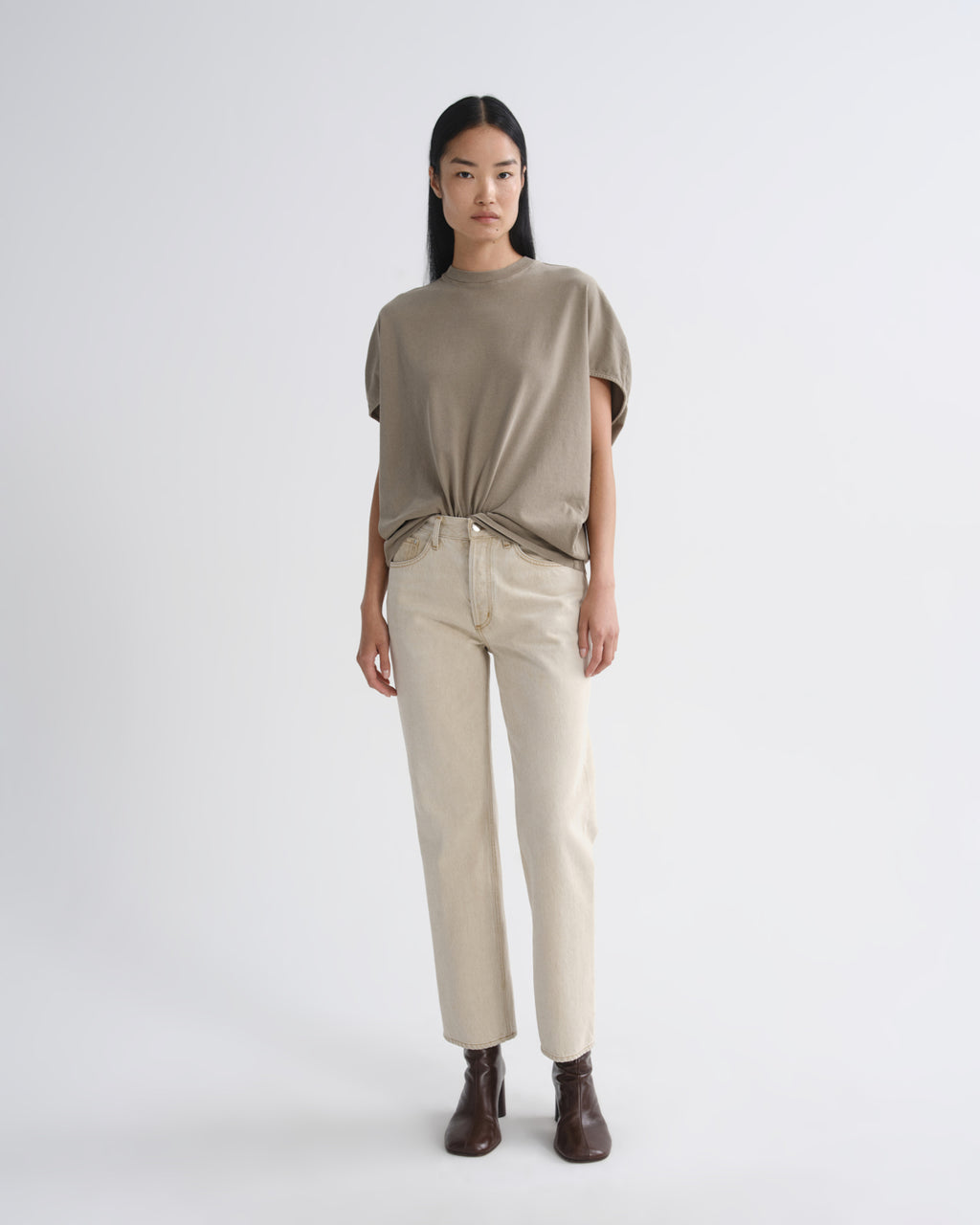 A model stands against a light gray studio background wearing a loose taupe short-sleeve top tied at the waist, straight beige pants, and dark brown ankle boots.