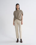 A model stands against a light gray studio background wearing a loose taupe short-sleeve top tied at the waist, straight beige pants, and dark brown ankle boots.