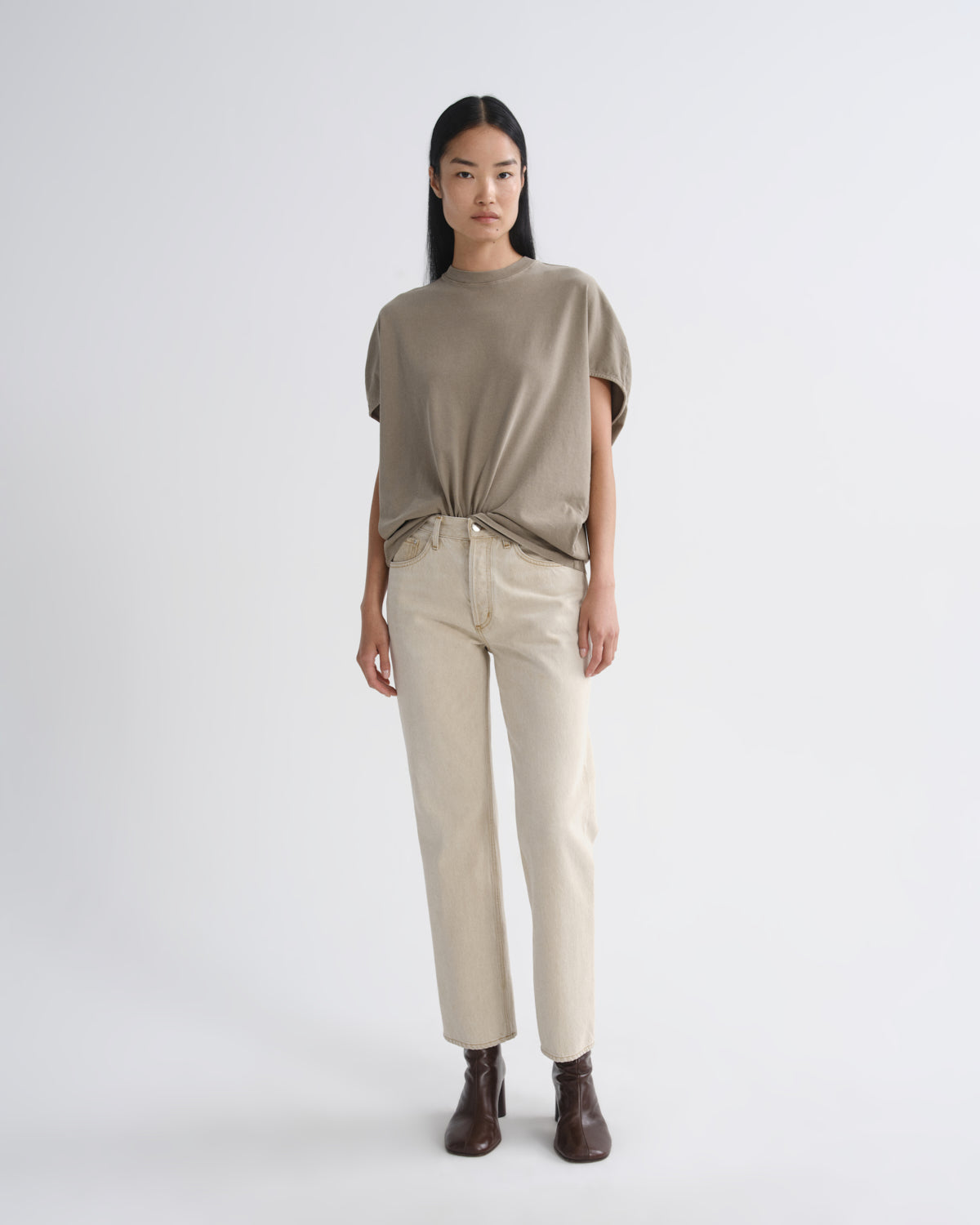 A model stands against a light gray studio background wearing a loose taupe short-sleeve top tied at the waist, straight beige pants, and dark brown ankle boots.