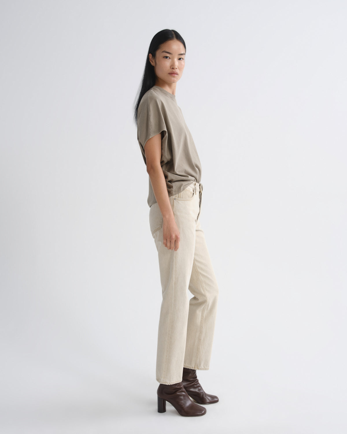 A model stands in profile wearing a loose taupe short-sleeve top, light beige wide-leg trousers, and dark brown heeled ankle boots against a plain light gray studio background.