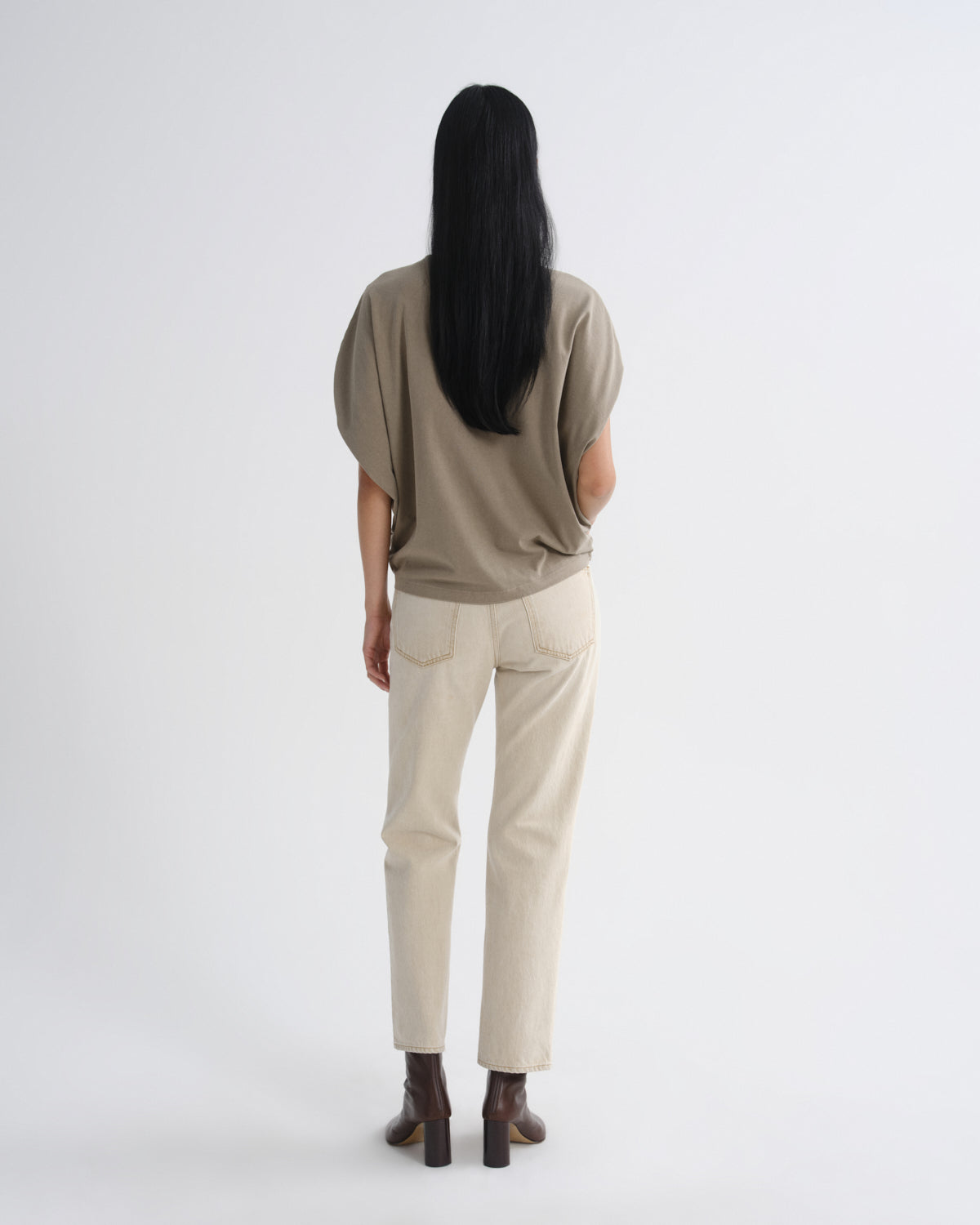 A woman viewed from behind wearing a loose taupe short-sleeve top, off-white straight-leg jeans, and dark brown ankle boots against a plain light gray studio background.