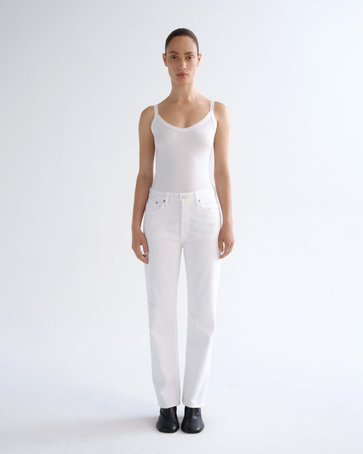A model stands against a seamless white studio background wearing a white ribbed camisole top, white straight-leg jeans, and black shoes.