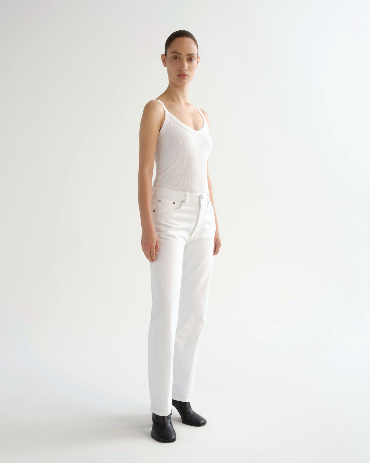 A model stands in a studio wearing a white ribbed camisole top, white high-waisted jeans, and black ankle boots against a plain white background.