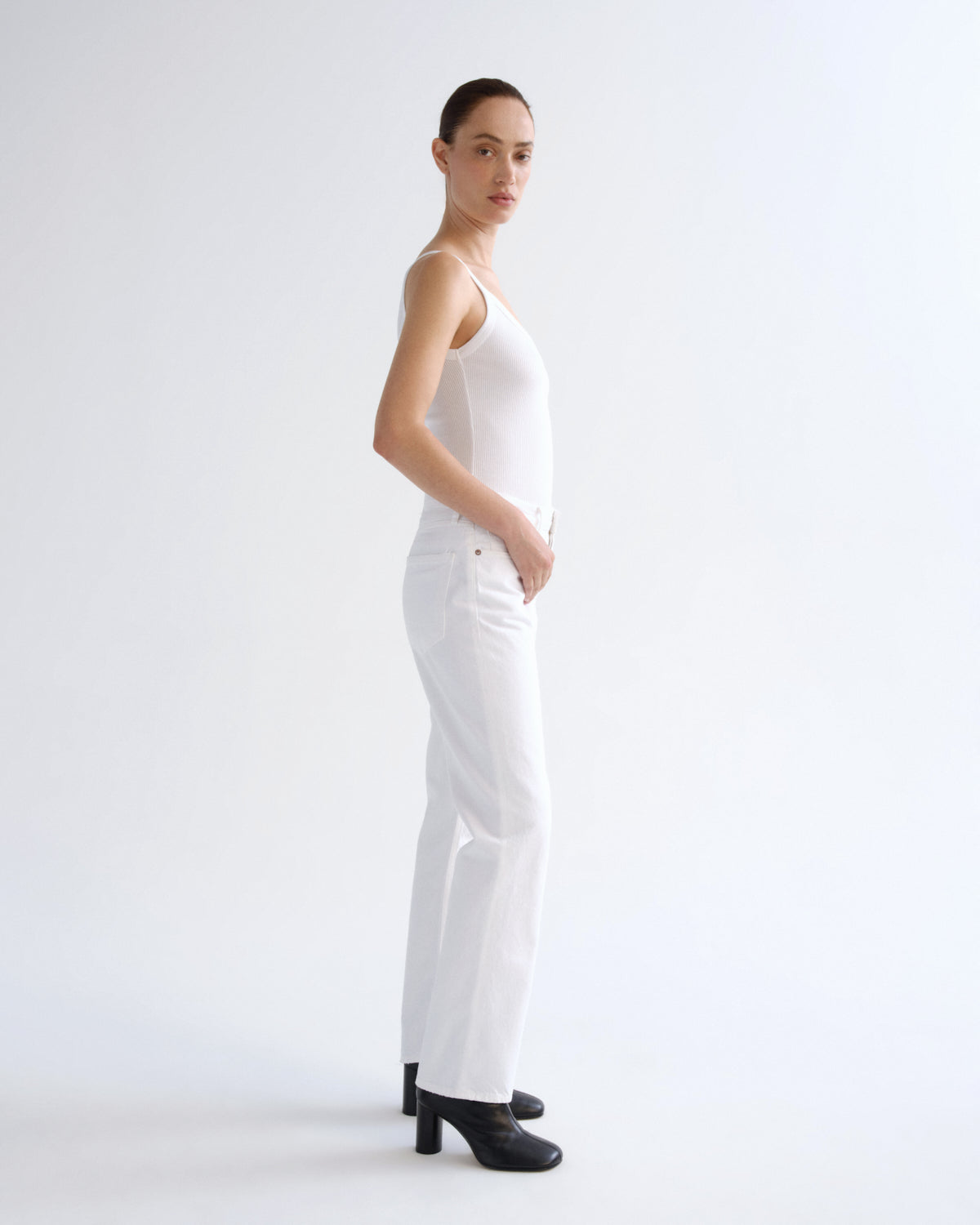 A woman stands in profile wearing a white sleeveless top and white pants with black high heels against a plain white studio background.