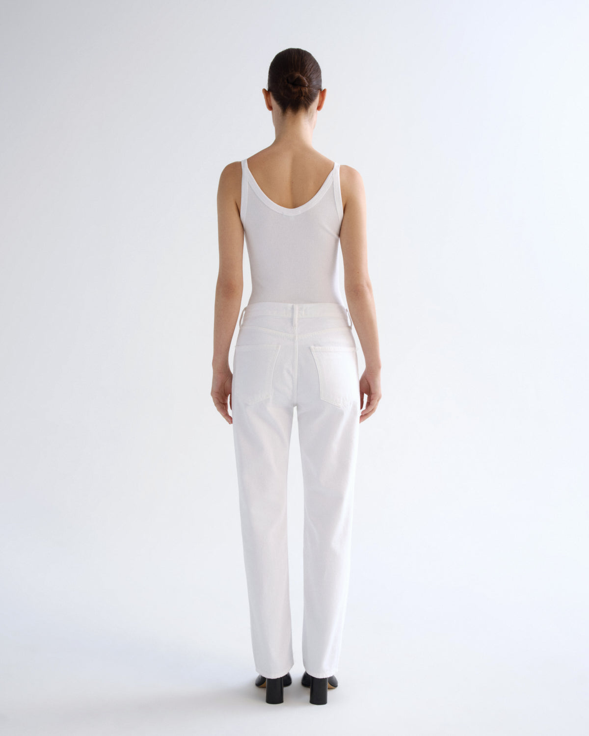 A model viewed from behind wearing a white sleeveless top and white high-waisted straight-leg jeans, paired with black block heels, against a plain white studio background.