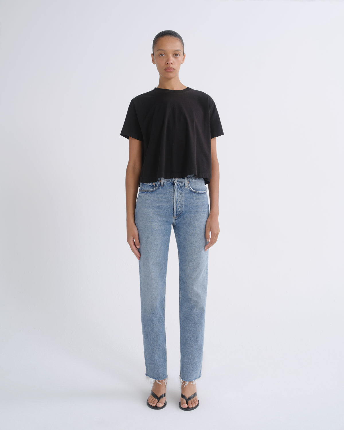 A model stands against a light gray studio background wearing a black short-sleeve crewneck t-shirt, light-wash blue straight-leg jeans, and black sandals.