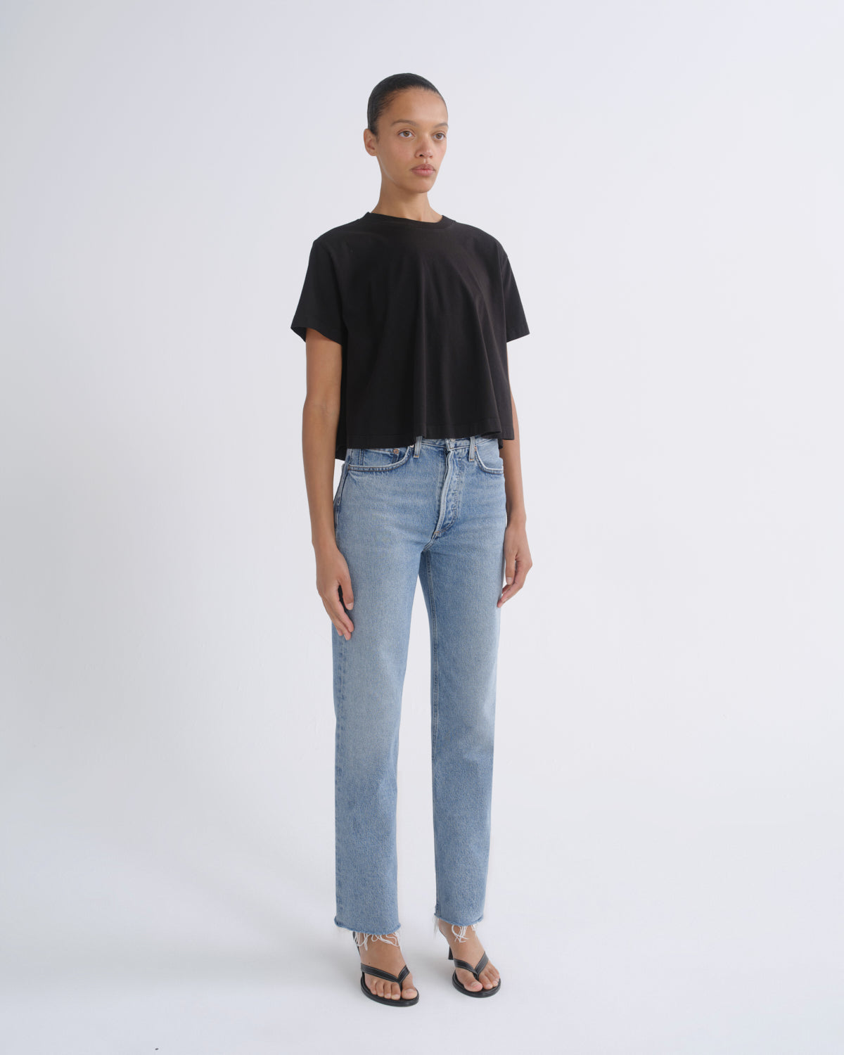 A model stands against a light gray studio background wearing a black short-sleeve cropped t-shirt, light-wash blue straight-leg jeans, and black thong sandals.