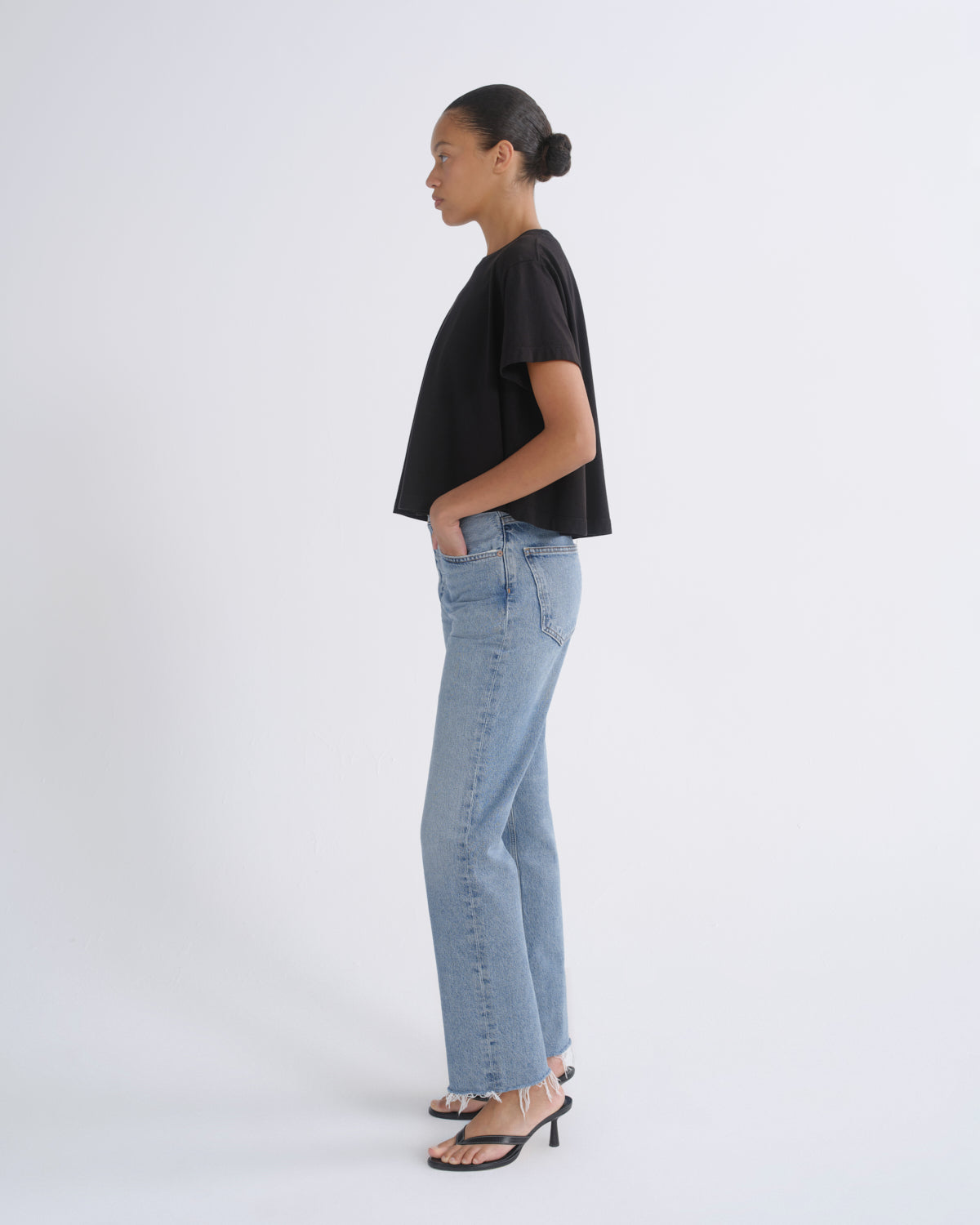 A woman in profile wearing a black cropped short-sleeve t-shirt, light-wash blue straight-leg jeans, and black heeled sandals, standing against a plain light gray studio background.