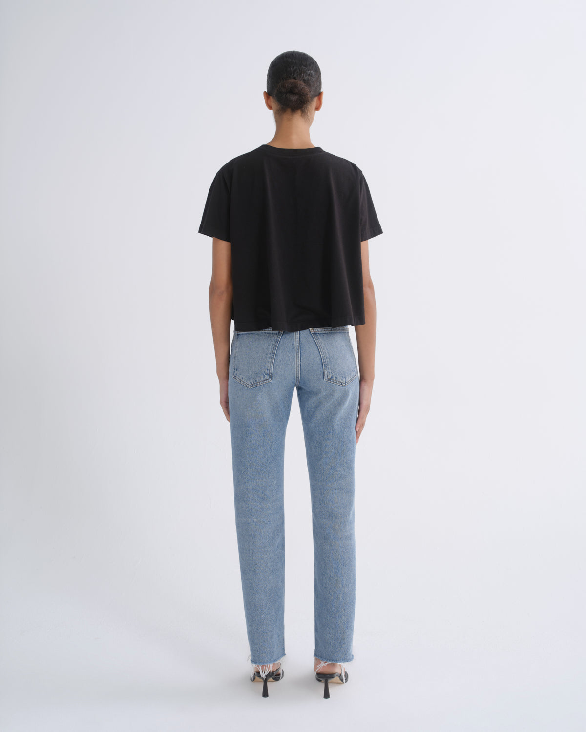 A model viewed from the back wearing a black short-sleeve crop top, light-wash blue jeans, and black high heels against a plain light gray studio background.