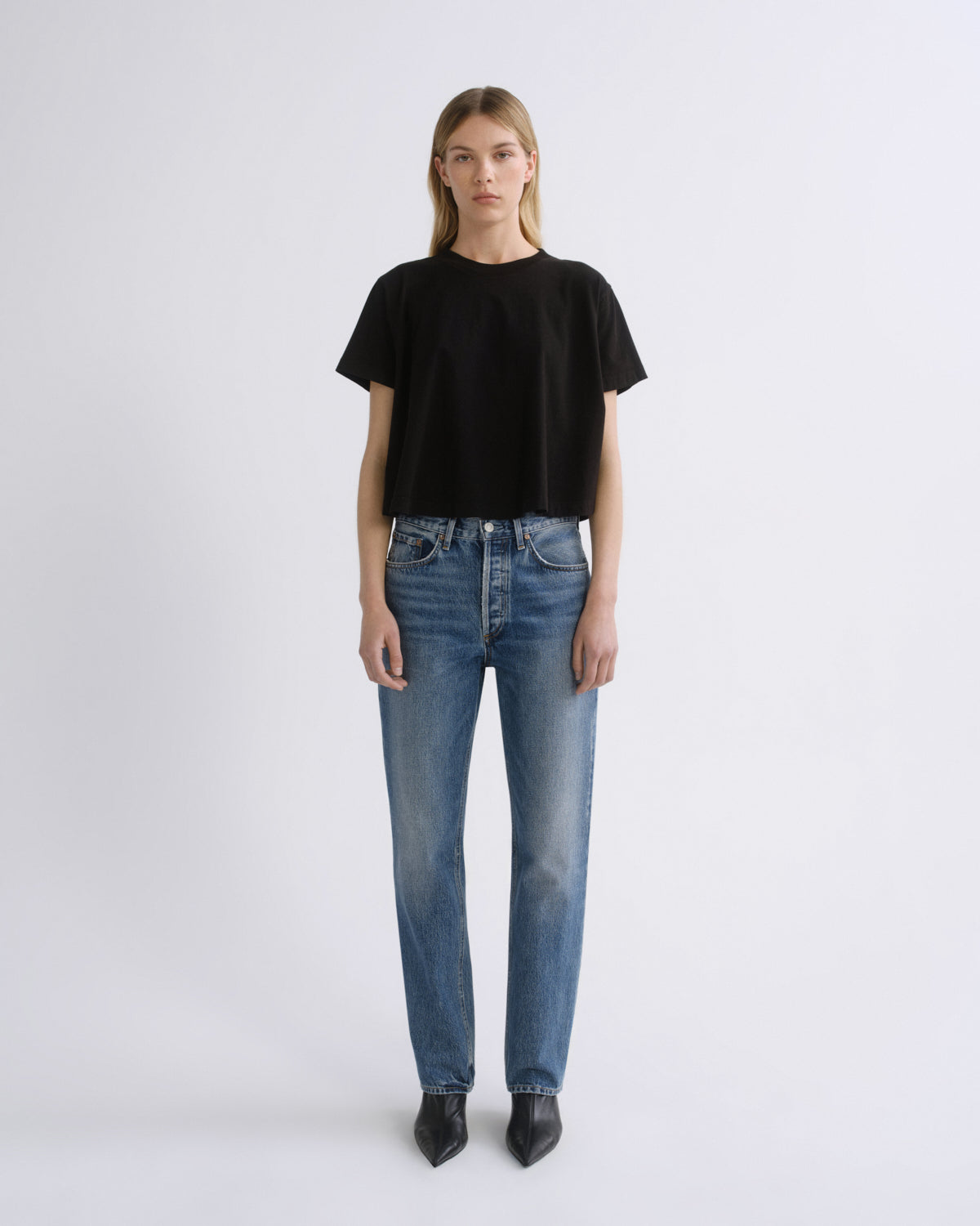 A model stands against a light gray studio background wearing a black short-sleeve crewneck t-shirt, blue straight-leg jeans, and black pointed-toe boots.