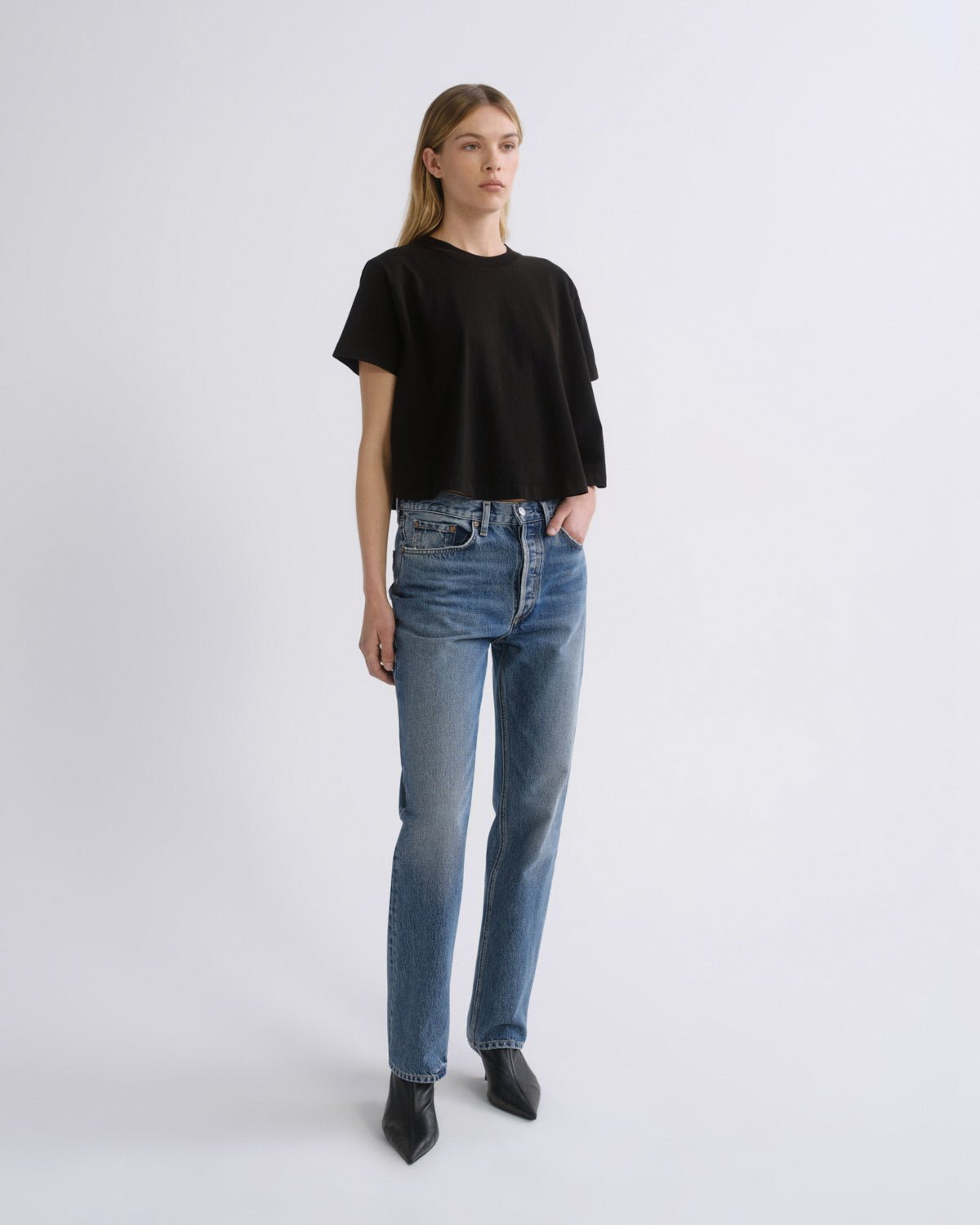A woman modeling a black short-sleeve cropped t-shirt and blue straight-leg jeans against a plain light gray studio background.